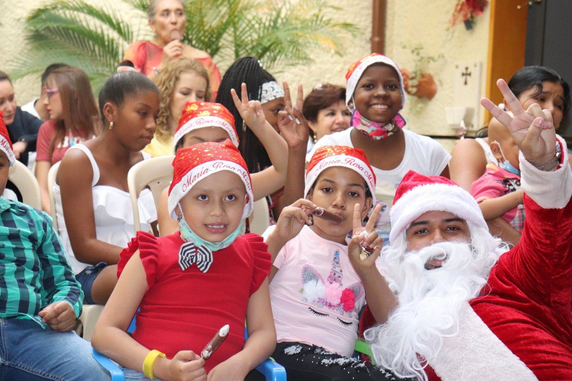 Niños con gorros de Papá Noel con Santa Claus en un evento, muchos sonriendo y haciendo señales de paz.