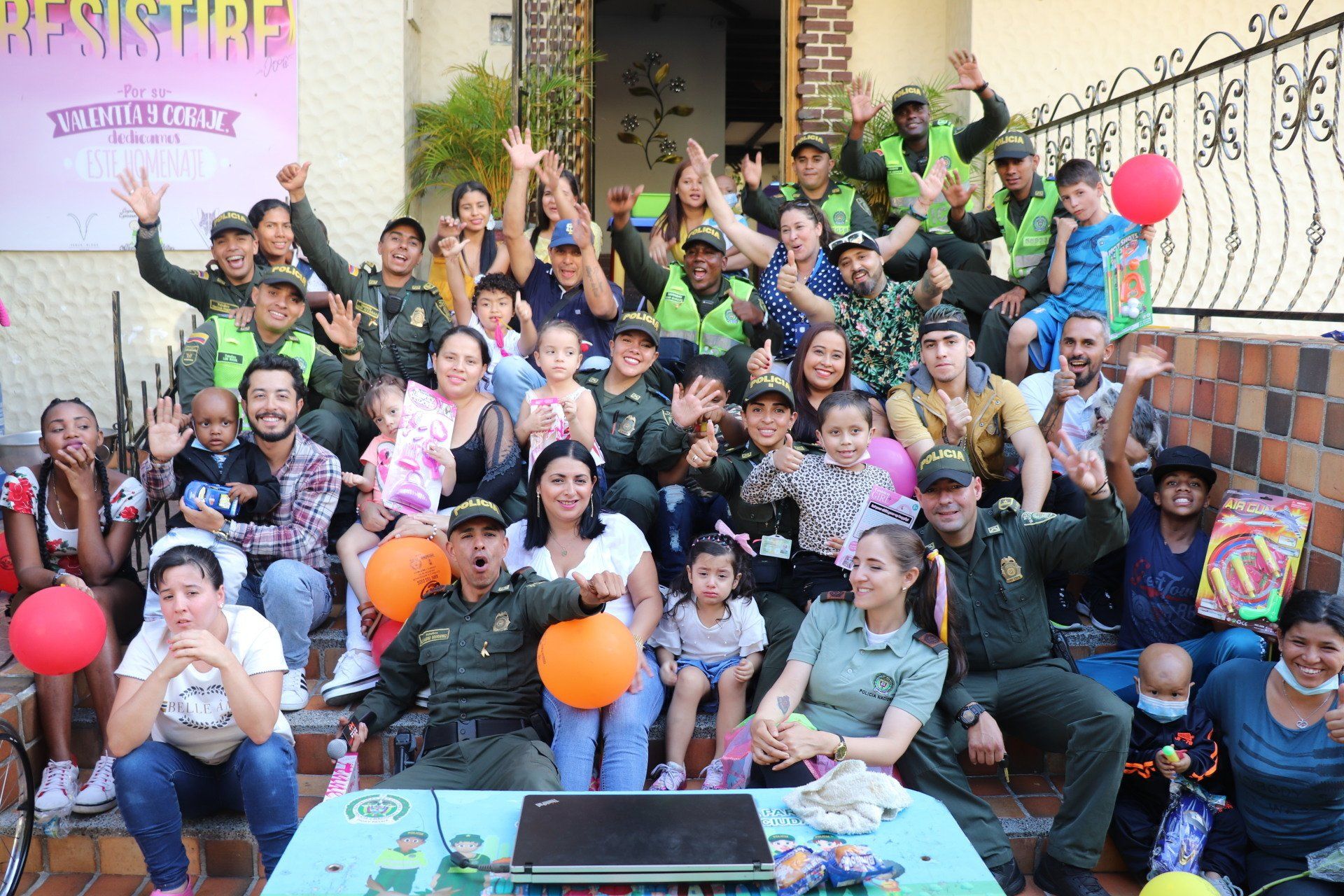 Un grupo de policías y civiles, incluyendo niños, sonríen y saludan al aire libre. Hay globos.