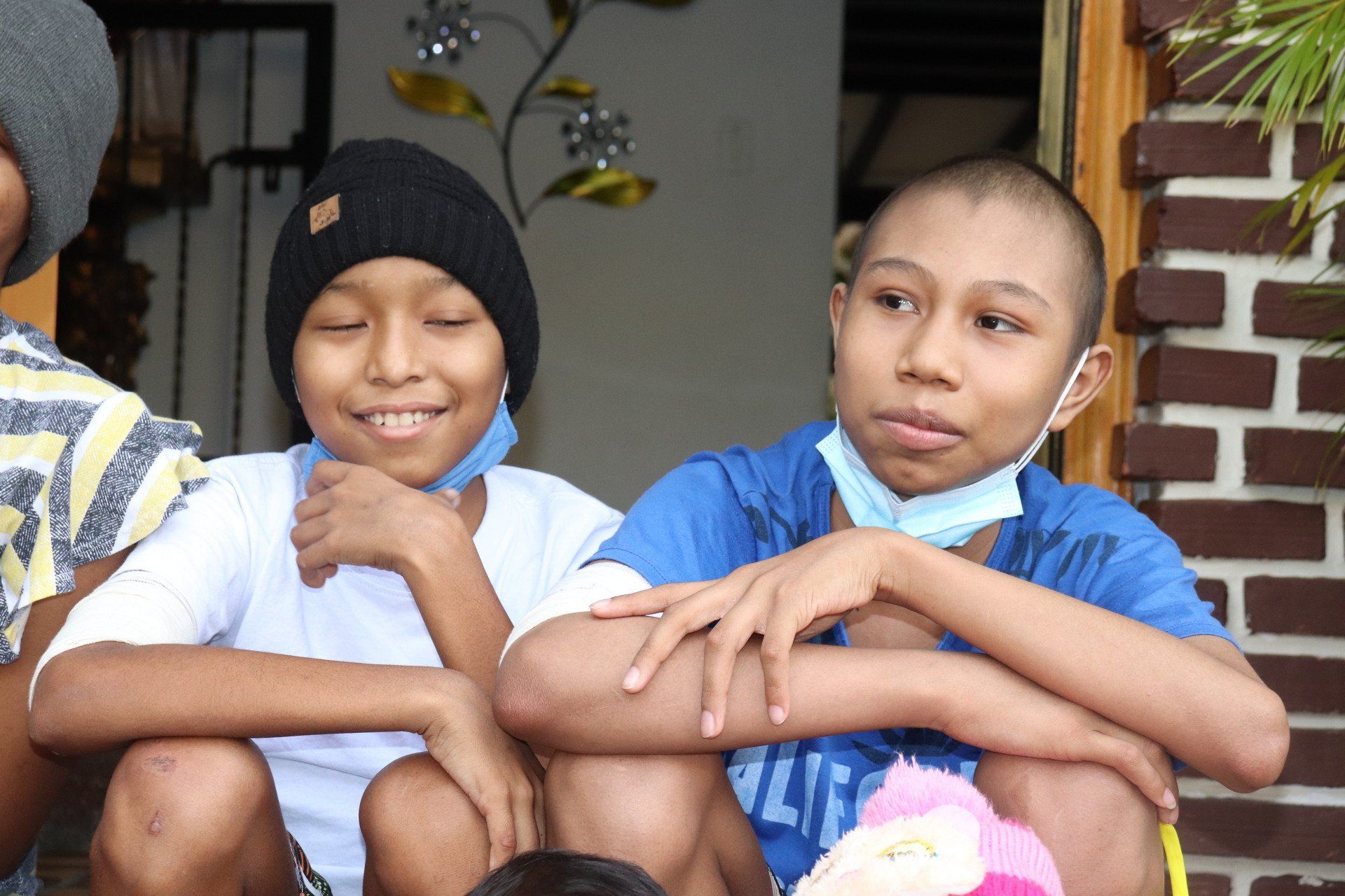 Dos chicos con la cabeza rapada y mascarillas, uno de ellos con gorro. Están sentados juntos, sonriendo.