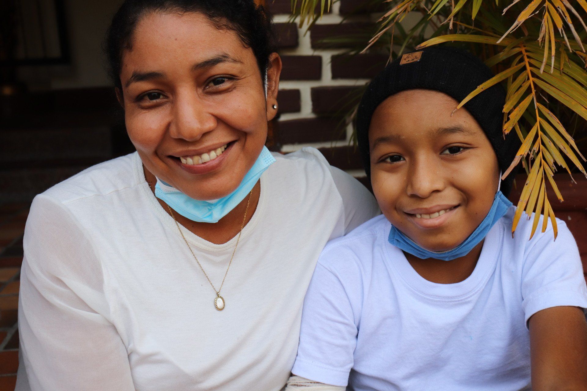 Mujer y niño sonriendo, con máscaras debajo de la barbilla, frente a una pared de ladrillos y una planta verde.