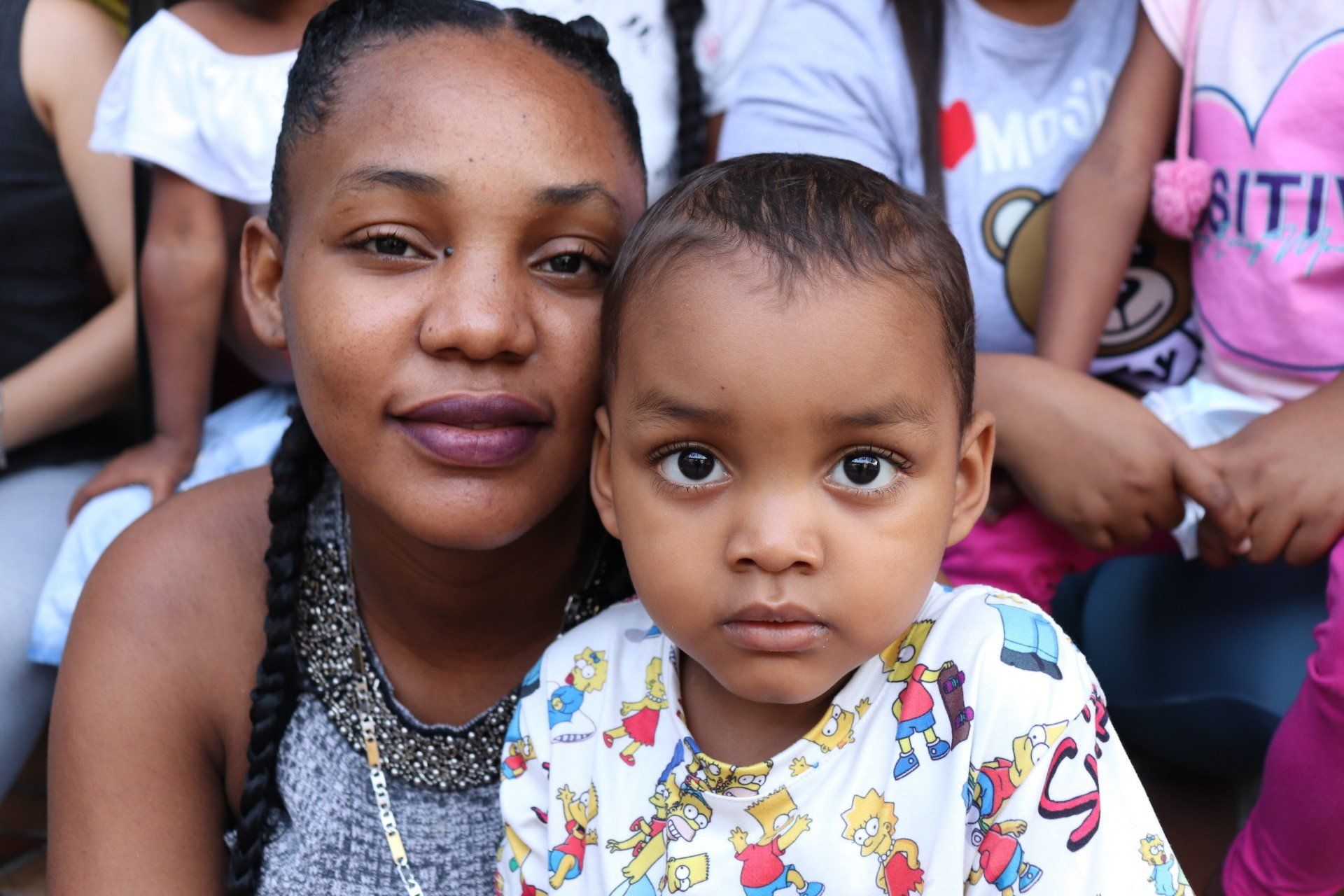 Mujer y niño mirando a la cámara. La mujer tiene piel oscura y trenzas. El niño tiene piel clara y camisa estampada.