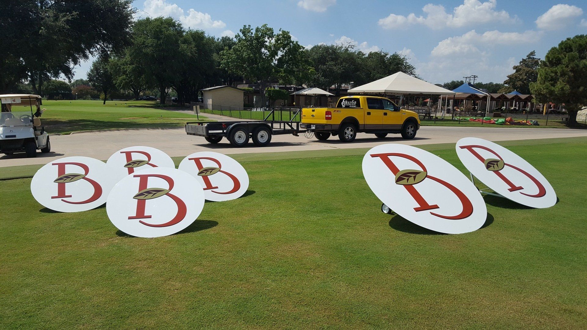 A yellow truck is towing a golf cart with bb signs on the grass