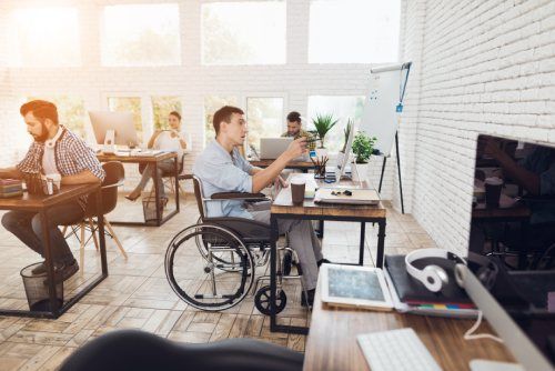 Ufficio con persone che lavorano alla scrivania, una delle quali su sedia a rotelle. Spazio luminoso e moderno con ampie finestre.