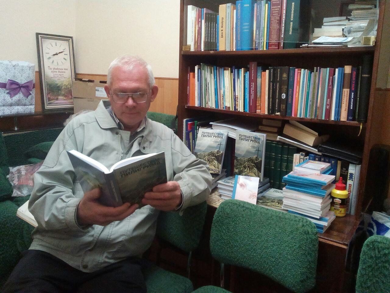 Een man met een bril leest een boek, zittend in een kamer met een boekenkast en een klok.