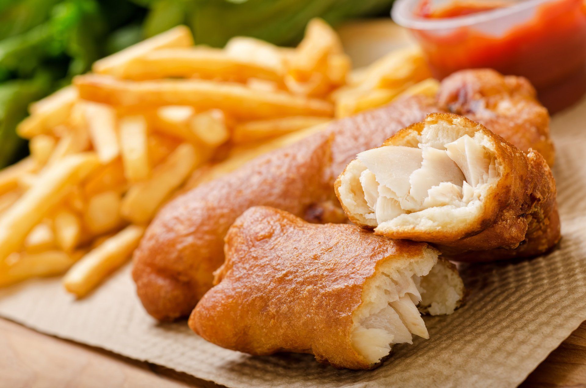 Fish and chips on a paper towel with a small container of ketchup and a side of green herbs.