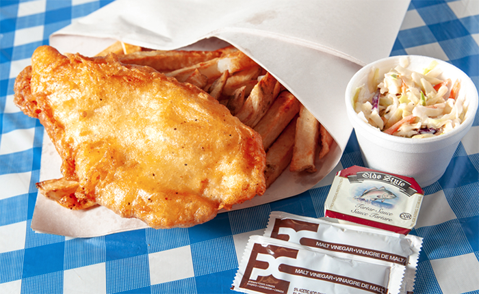 Fish and chips with coleslaw and condiments on a blue and white checkered surface.
