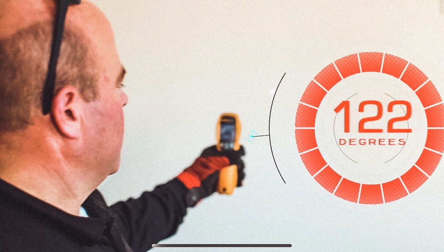 Man uses temperature gun on a wall, displaying 122 degrees. Orange glove and circle display.