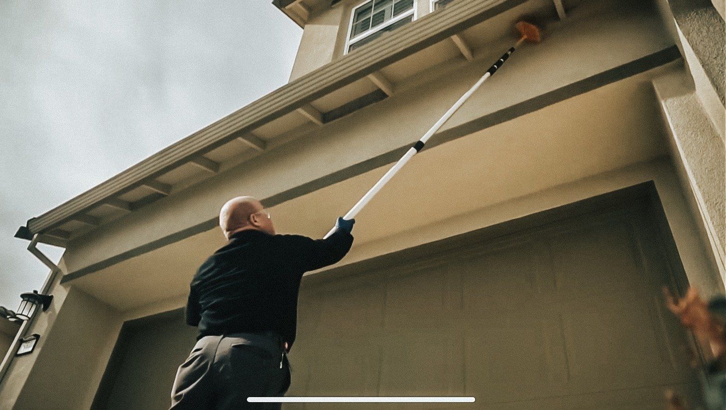 Man cleaning the eaves of a house with a long-handled brush, exterior shot.
