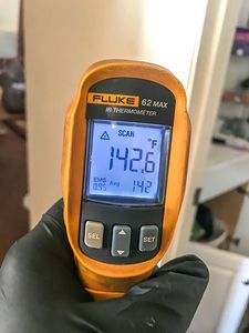 Hand holding an orange Fluke 62 MAX infrared thermometer displaying a temperature of 142.6°F.