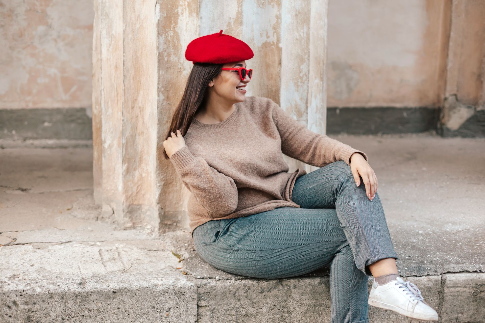 Una donna con un basco rosso e occhiali da sole sorride, seduta su una sporgenza di pietra, appoggiata a una colonna.