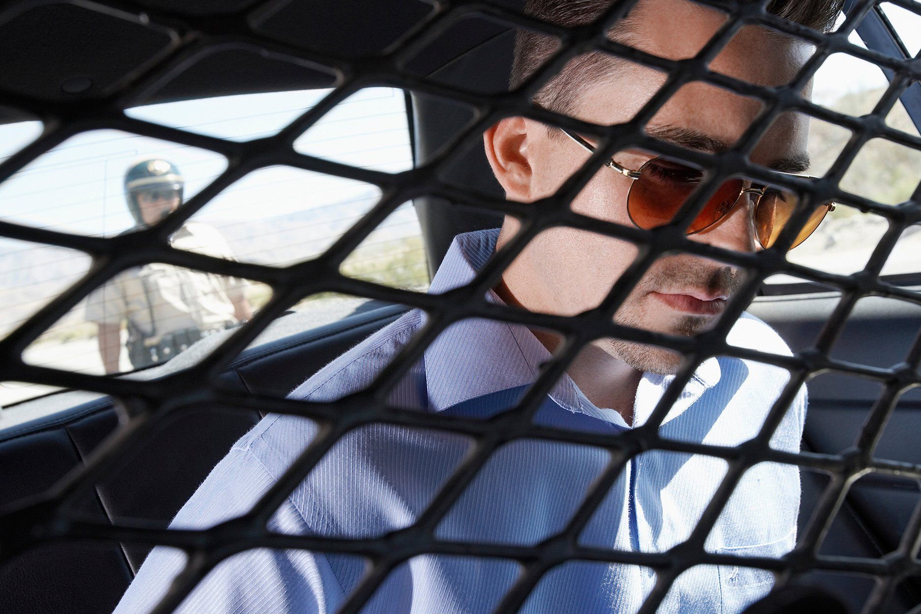 Man in sunglasses in back of police car, looking down. Policeman standing outside.