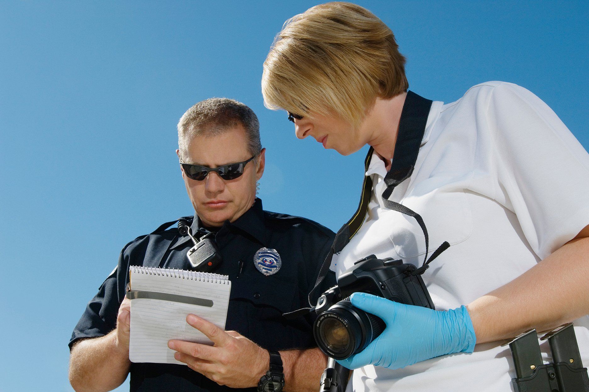 Police officer looking at a notepad with a photographer, who holds a camera. Bright blue sky.