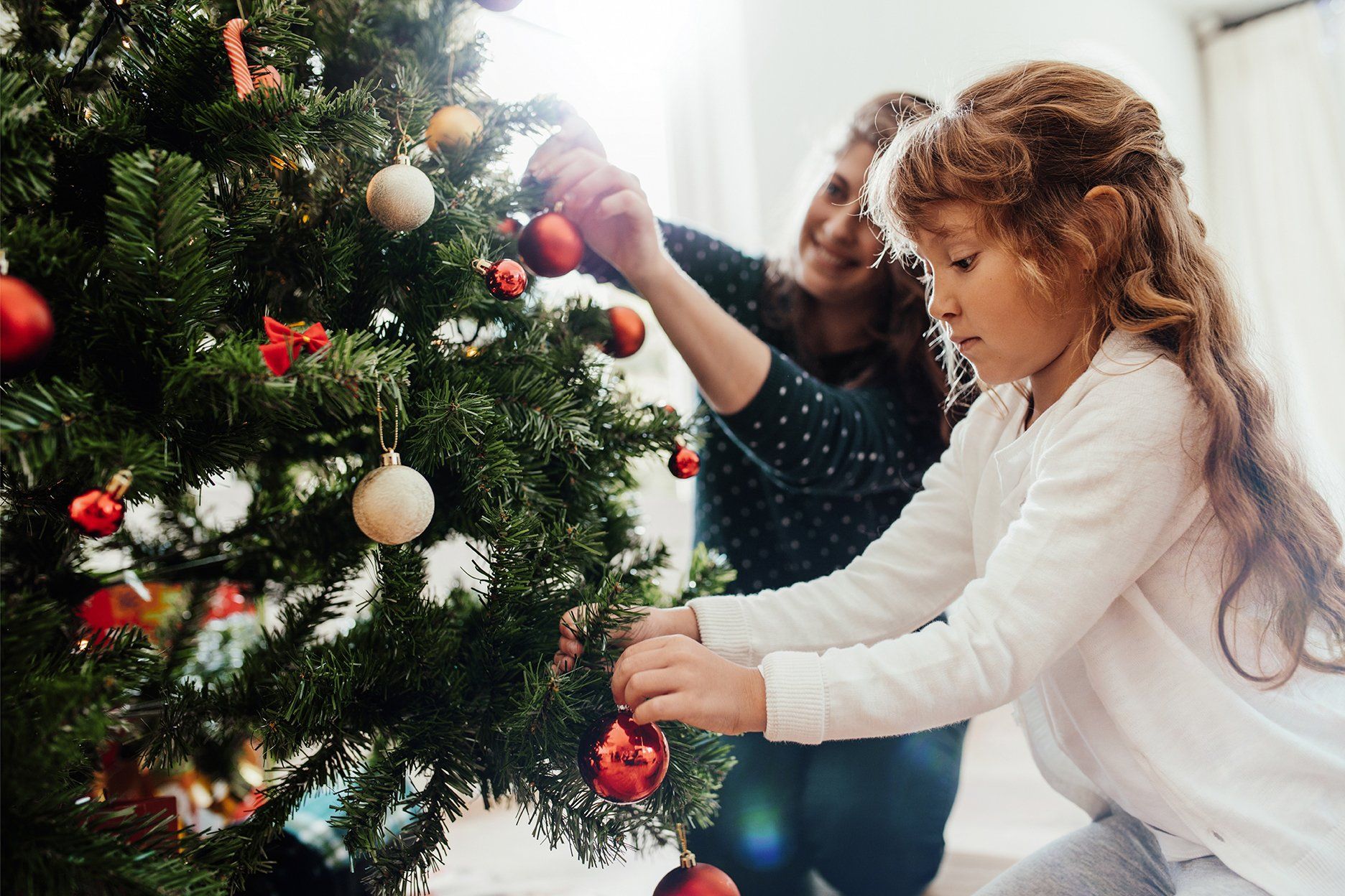 Woman and child decorating a Christmas tree with red and gold ornaments in a bright room.