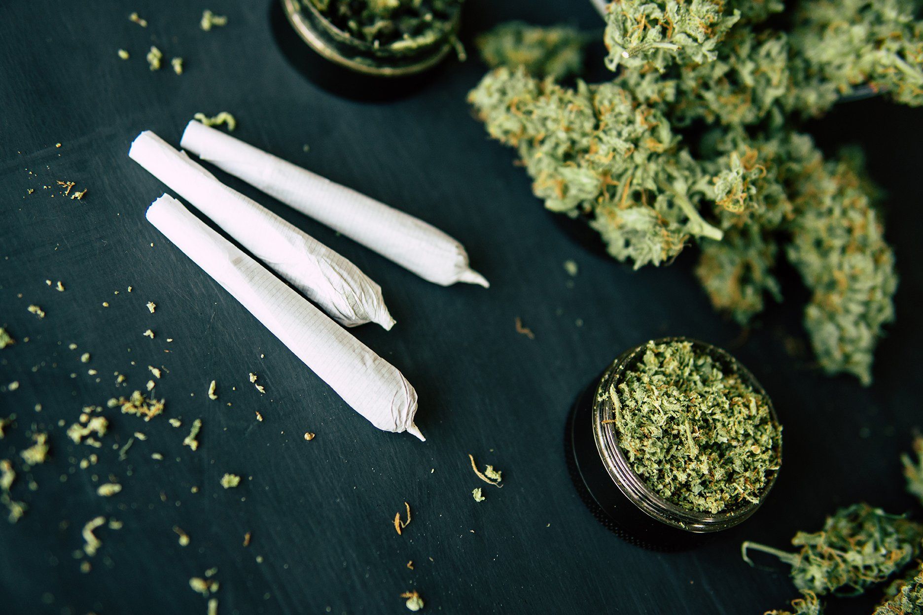 Three rolled joints and marijuana buds with a grinder on a dark surface.