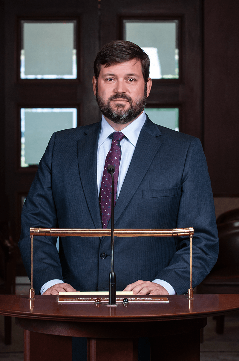 Man in a suit standing at a podium, looking forward. Dark blue suit, dark tie, formal setting.