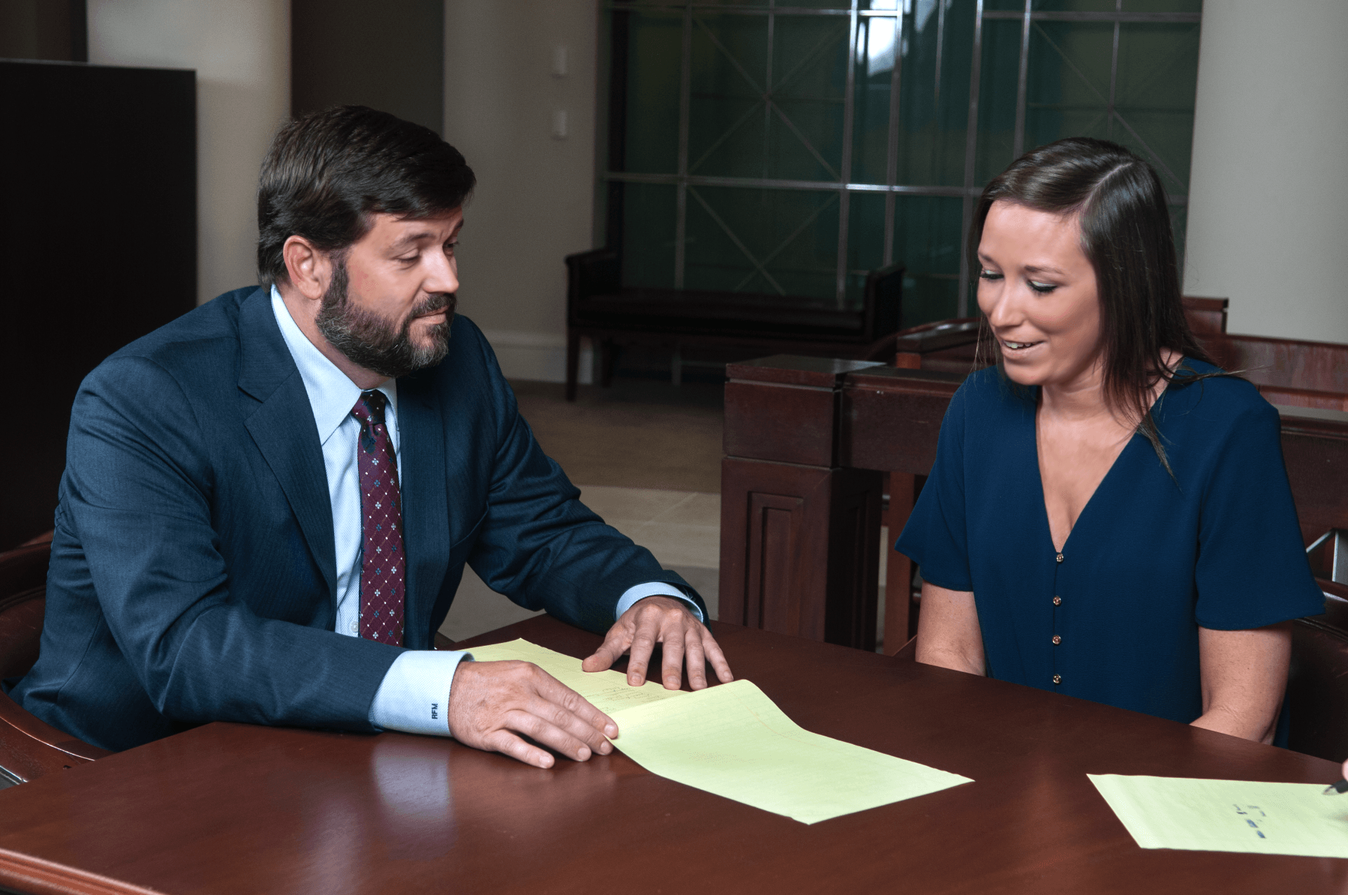 Man in suit reviews documents with woman in a dark blue dress at a table. Wooden desk, courtroom setting.