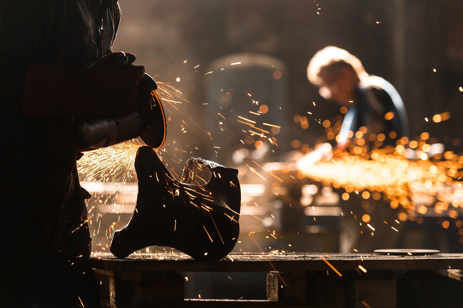 Person using a grinder, sparks flying. Another person works in the background in a workshop.