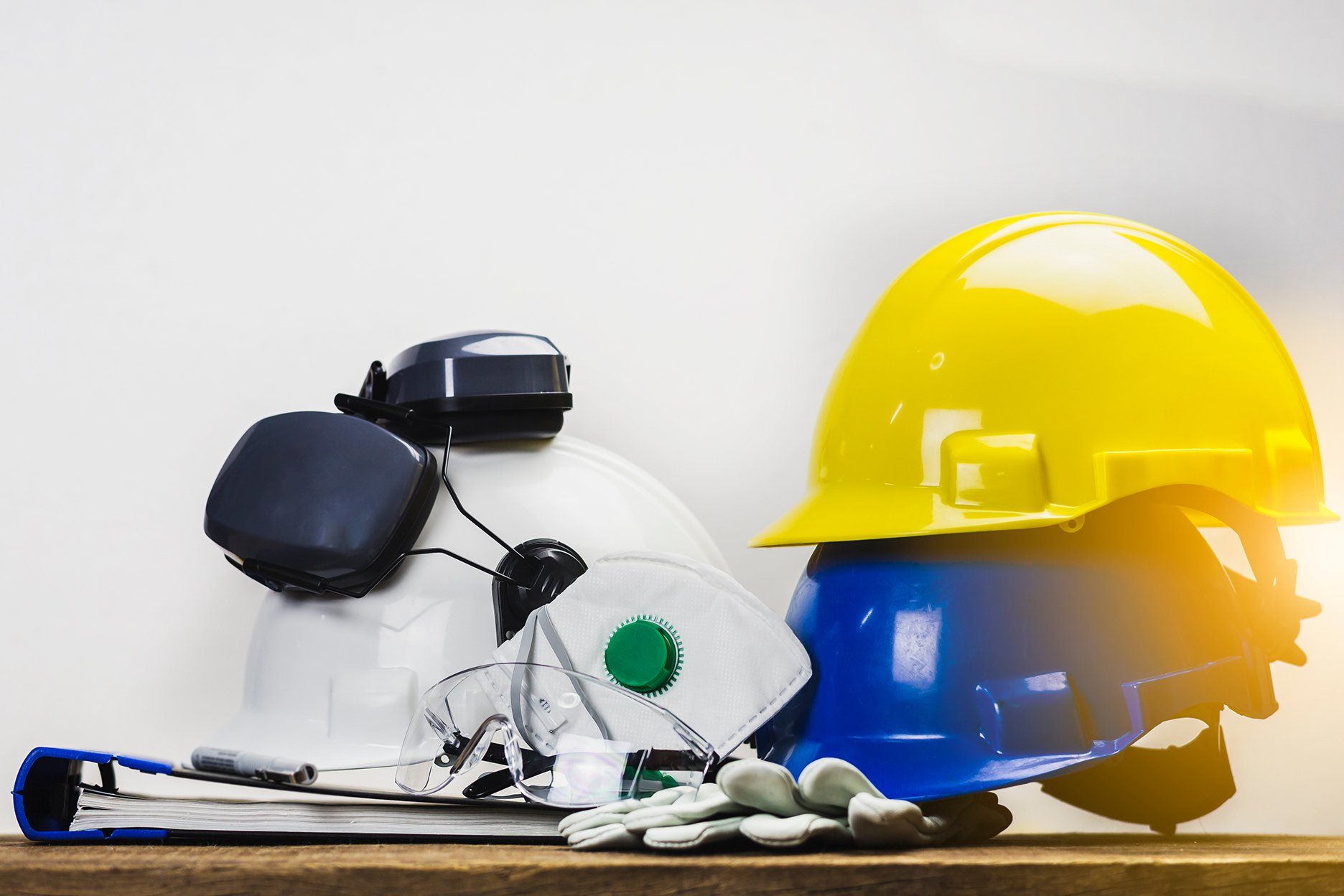 Pile of safety equipment: hard hats (yellow, blue, white), ear protectors, mask, gloves, and paperwork on a wooden surface.