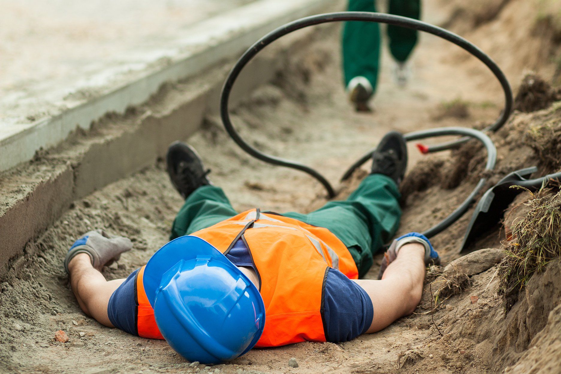 Construction worker lying on the ground in a trench, possibly injured, with a blue hard hat and orange vest.