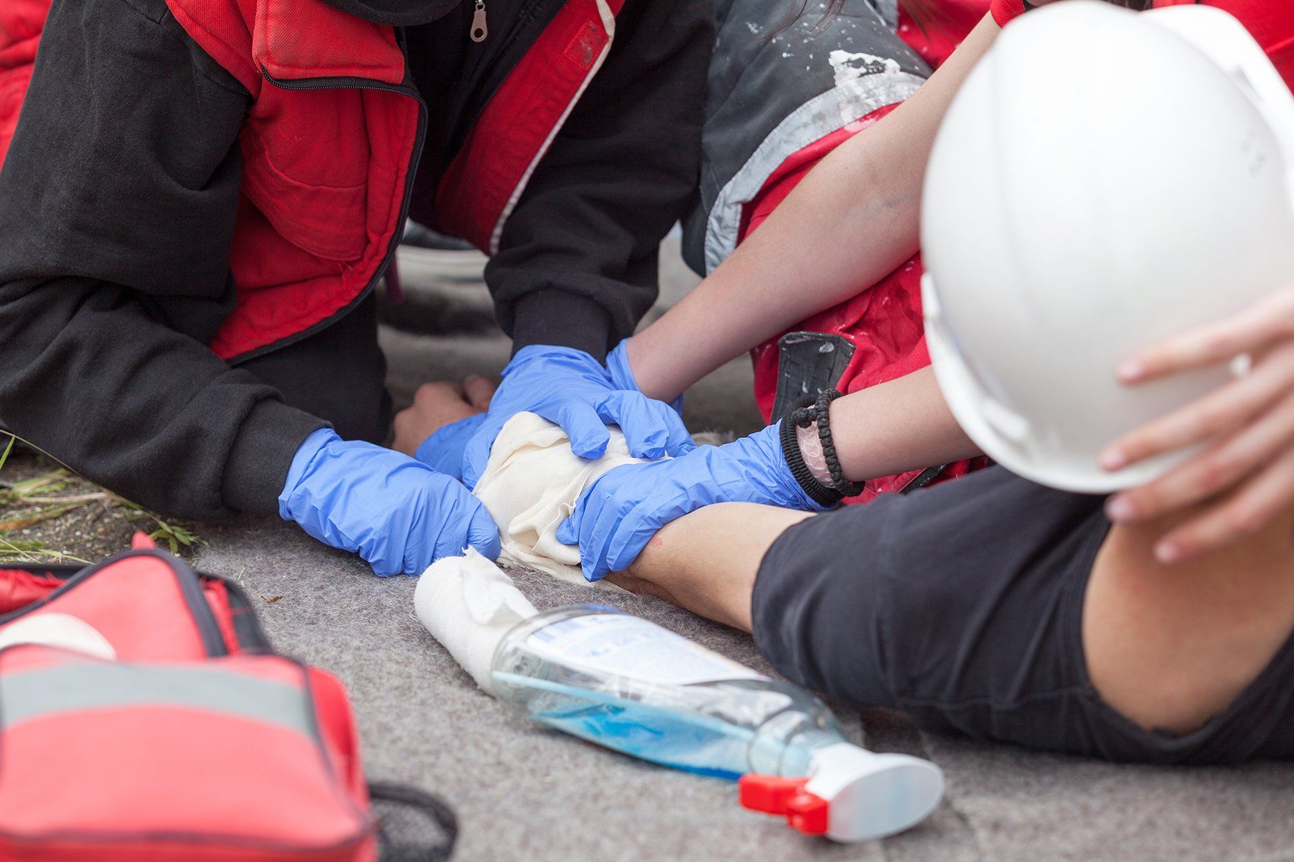 People treating an injured person with a white bandage on a gray surface; medical supplies nearby.