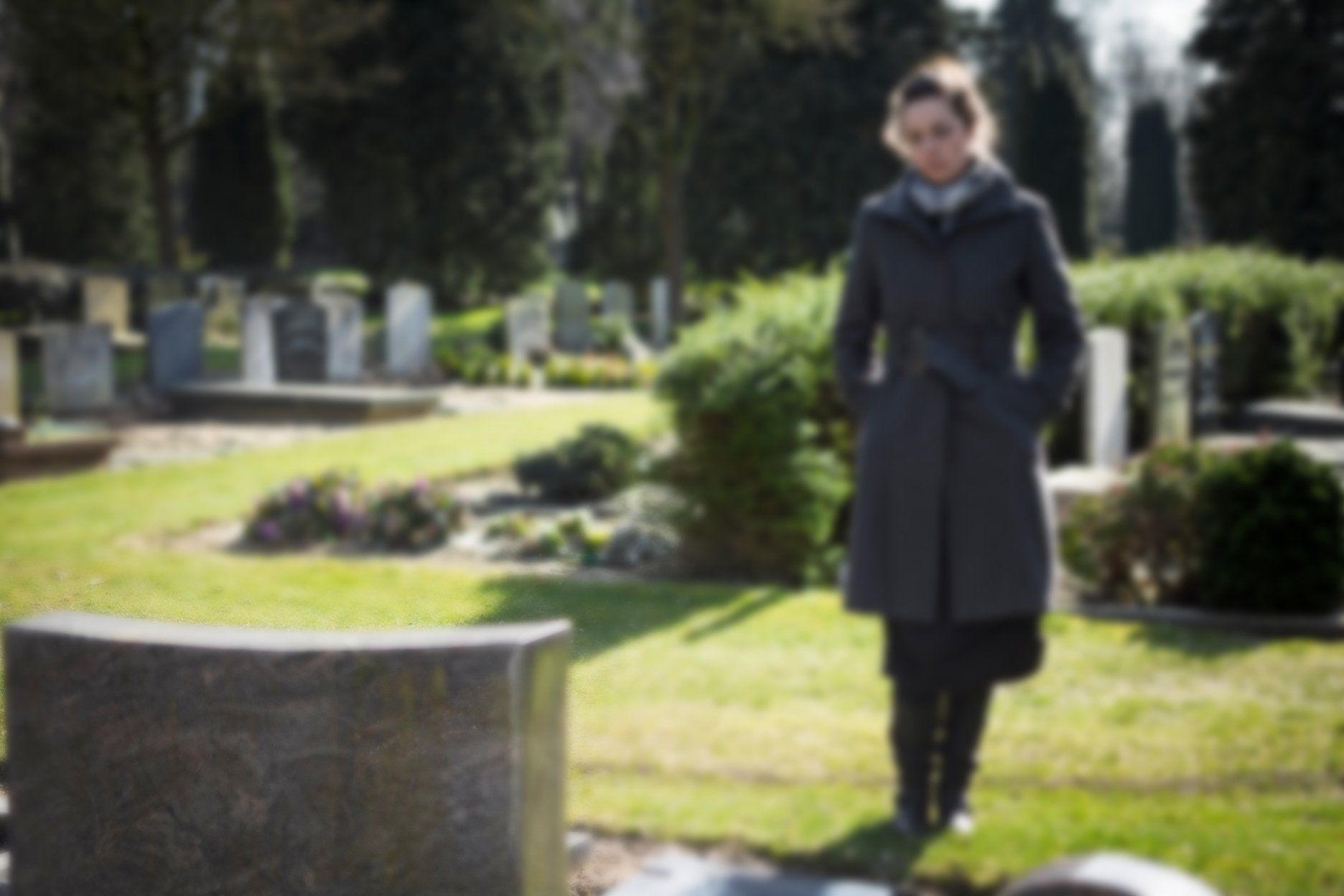 Woman in a gray coat stands by a headstone in a cemetery on a sunny day.