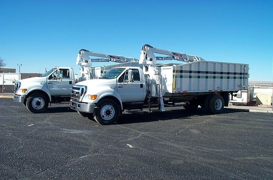 Two white work trucks with crane arms on a black asphalt lot under a clear blue sky.