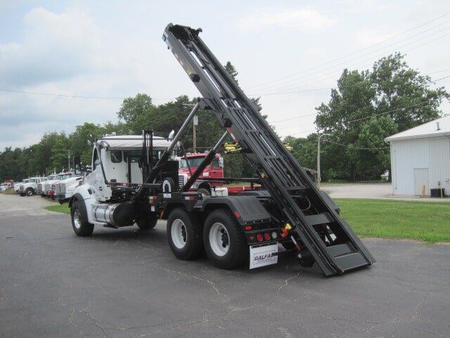 White roll-off truck with black lifting mechanism, parked outdoors, with trees and a building in the background.
