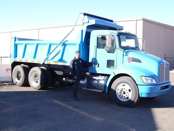 Blue dump truck with a person standing beside it on a sunny day.