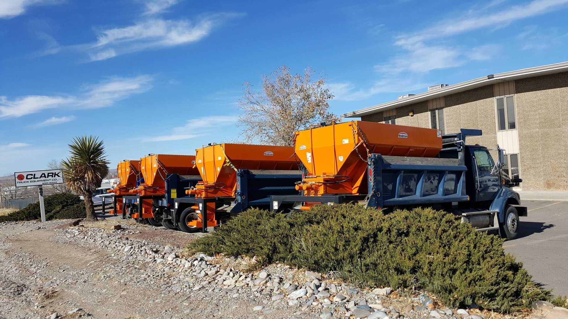 Four orange and blue salt trucks parked in a row on a sunny day.