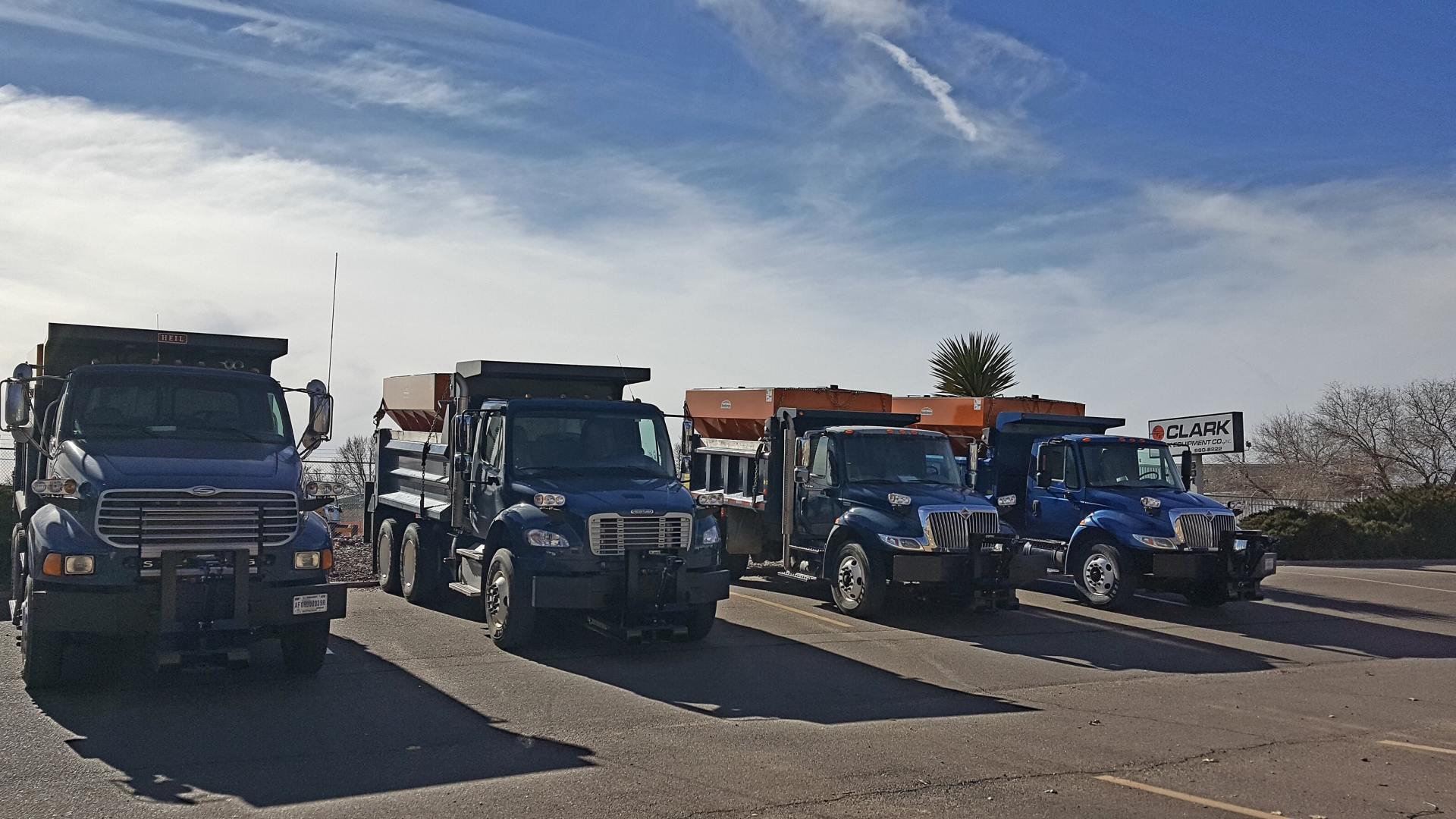 Four dark blue dump trucks parked in a row on a paved lot under a blue sky.