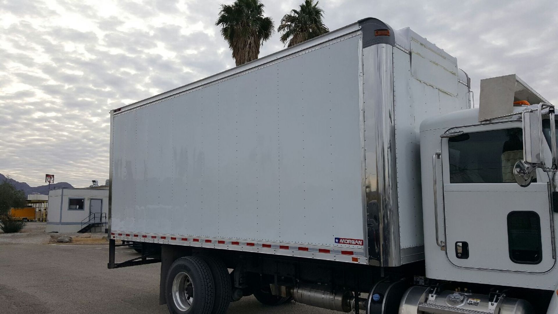 White box truck parked outdoors under a cloudy sky, near palm trees and a building.