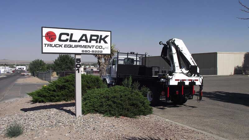 Clark Truck Equipment Co. sign, white truck with crane in front of building and bushes, sunny day.