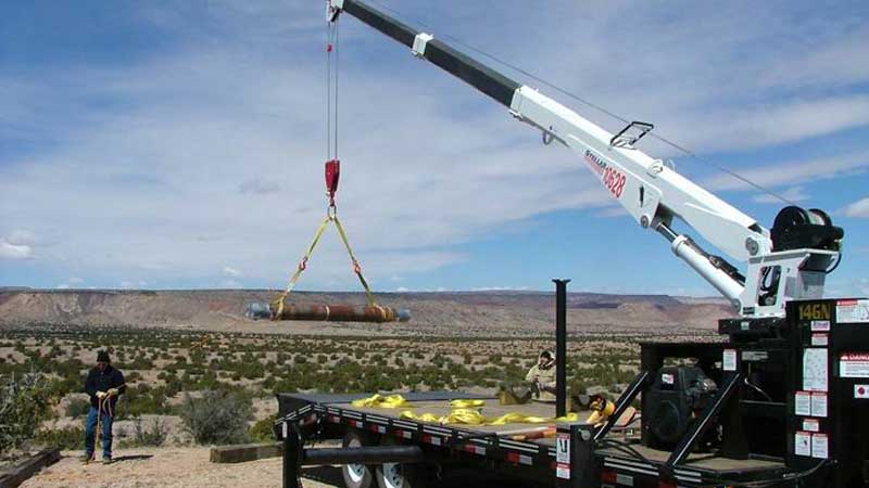 A crane lifting a large cylindrical object outdoors in a desert landscape; person watches.