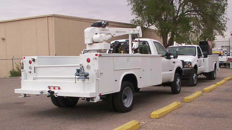 White utility truck with crane, towing another truck, parked in front of a building.
