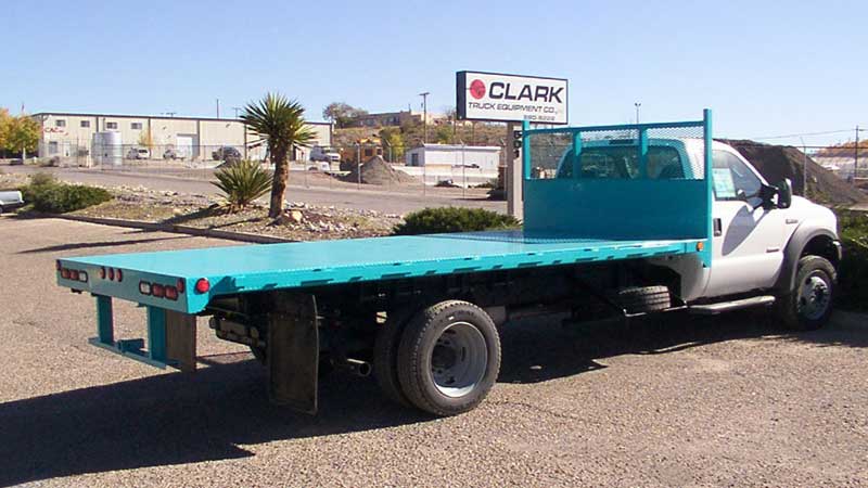 White flatbed truck with turquoise bed, parked on a sunny day in front of a business.