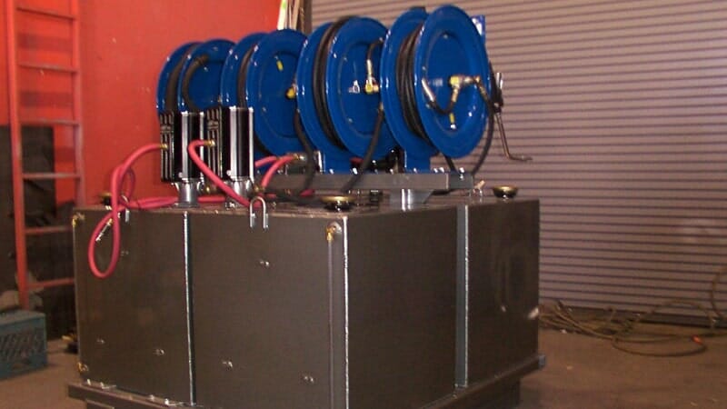 Metal tank with five blue hose reels mounted on top, set in a workshop.