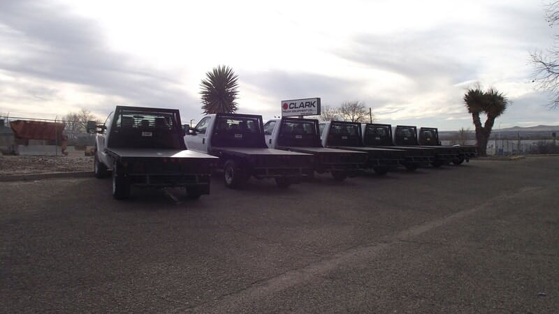 A line of flatbed trucks parked on asphalt under a cloudy sky; a sign is visible in the background.