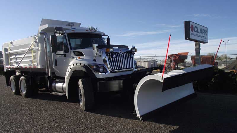 White snow plow truck on pavement, with a commercial building in the background.