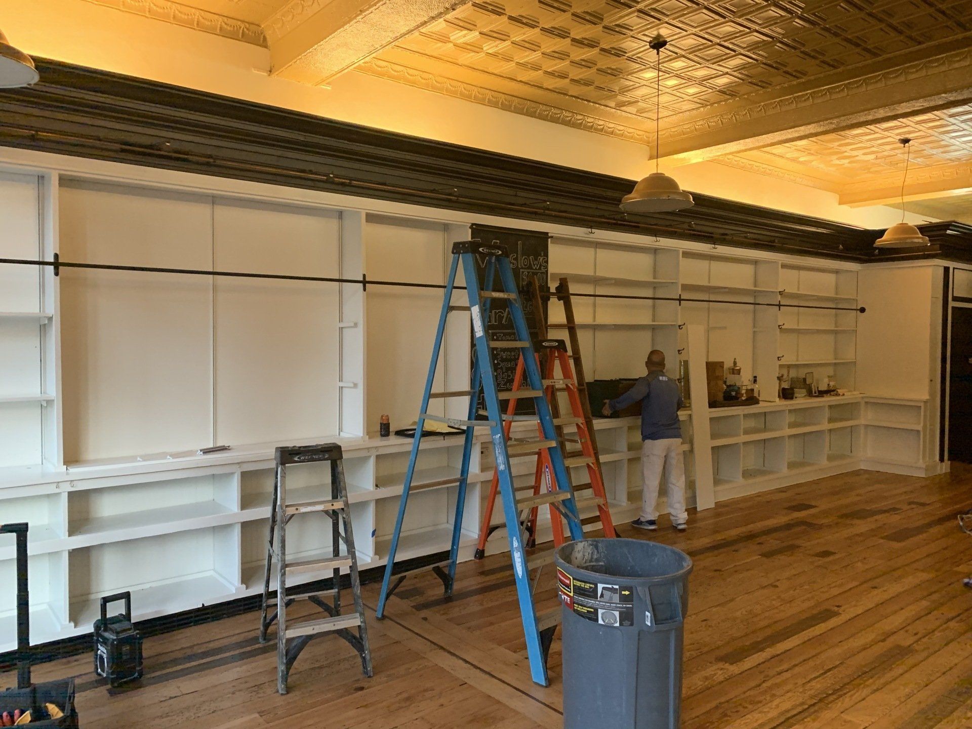 Workers installing shelving in a large room with ladders, tools, and unfinished wood flooring