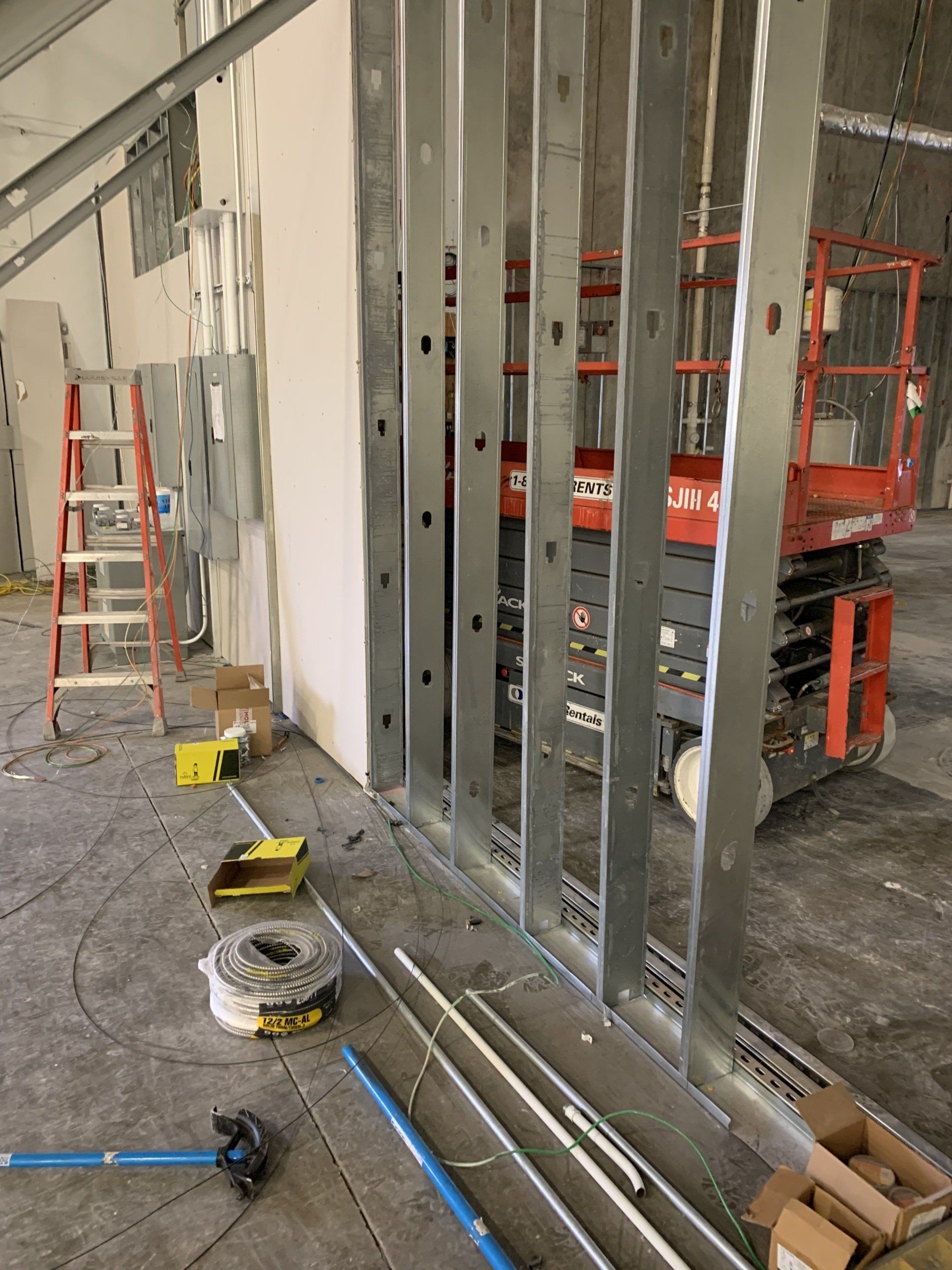 Interior construction site with metal wall studs, ladders, tools, and a scissor lift in the background