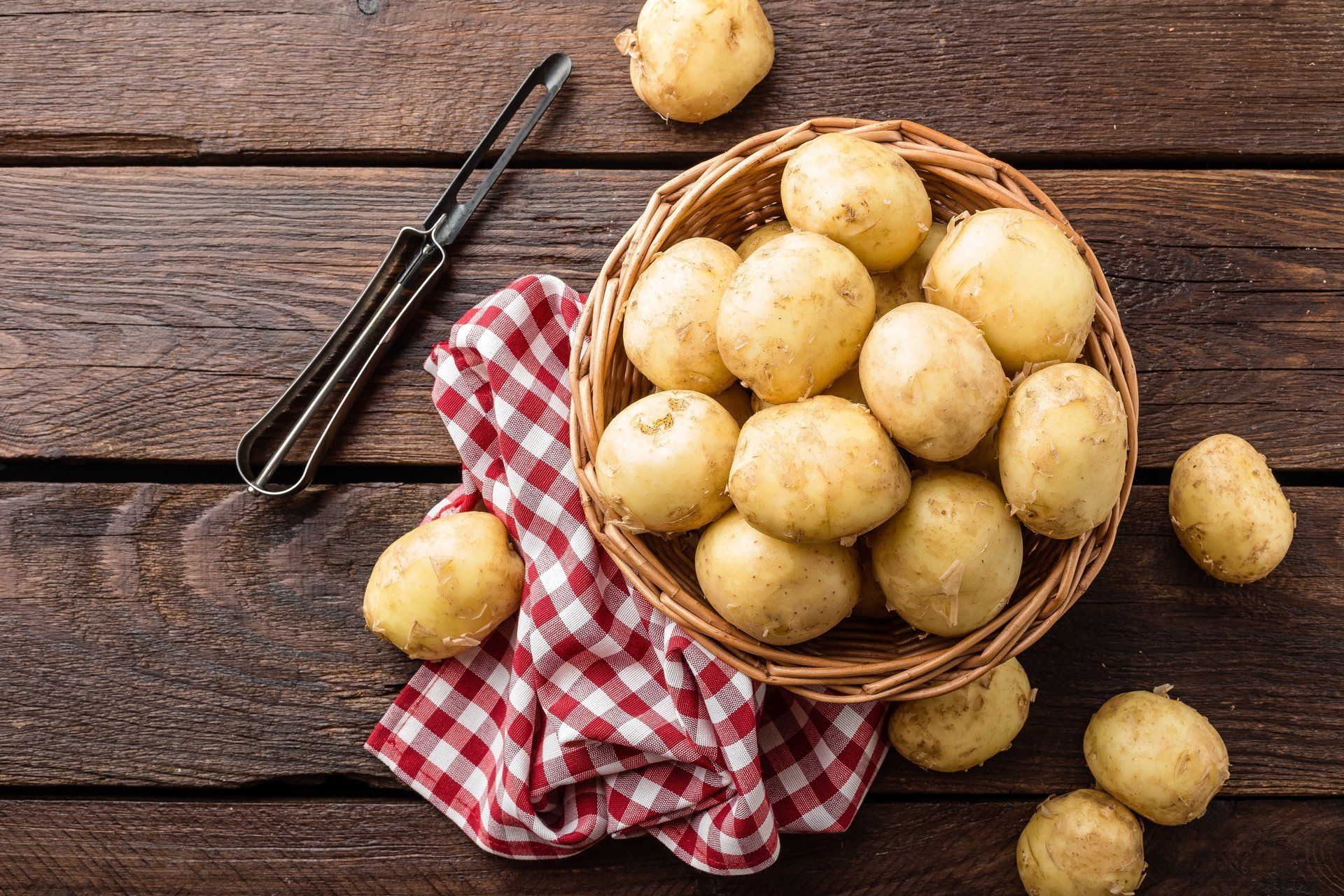 Auf einem dunklen Holztisch steht ein Korb mit kleinen, runden Kartoffeln neben einem Sparschäler und einem rot-weiß karie