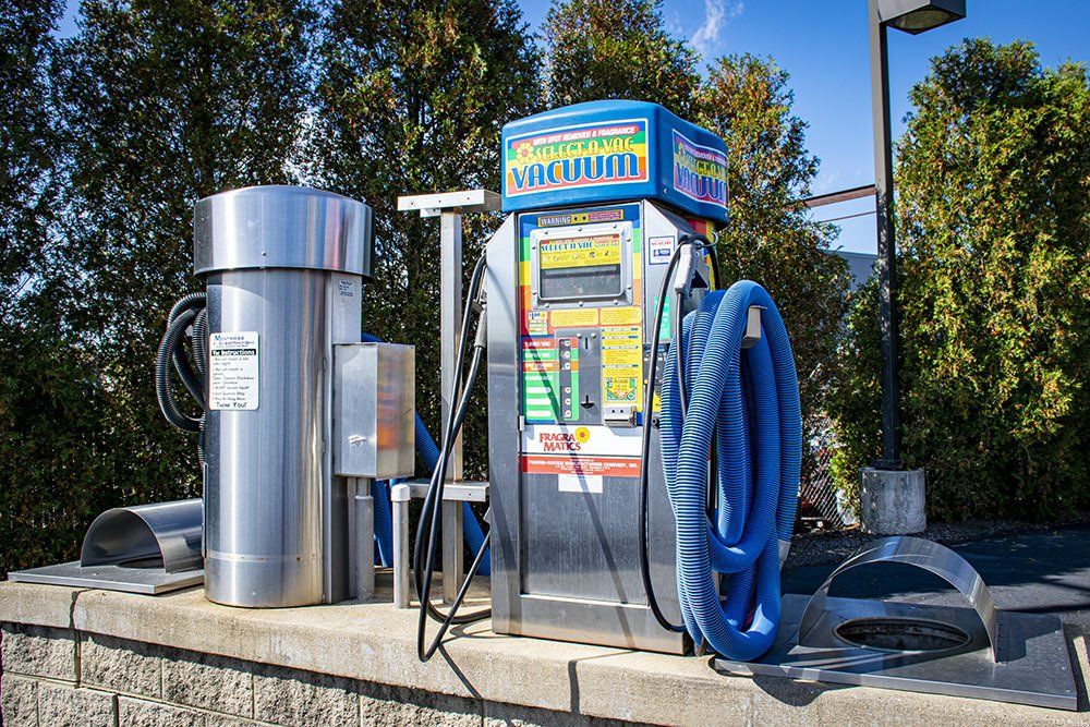 A car wash station with vacuum and hose against green trees.