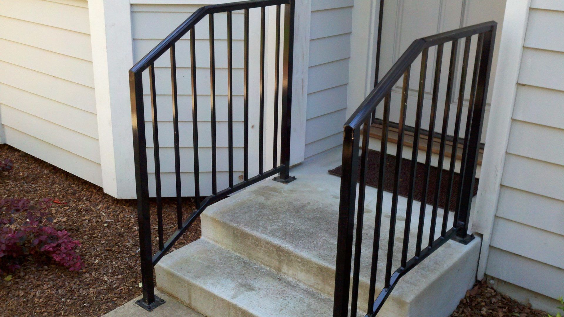Black metal handrails on a concrete staircase leading to a door. The setting is outdoors, daytime.