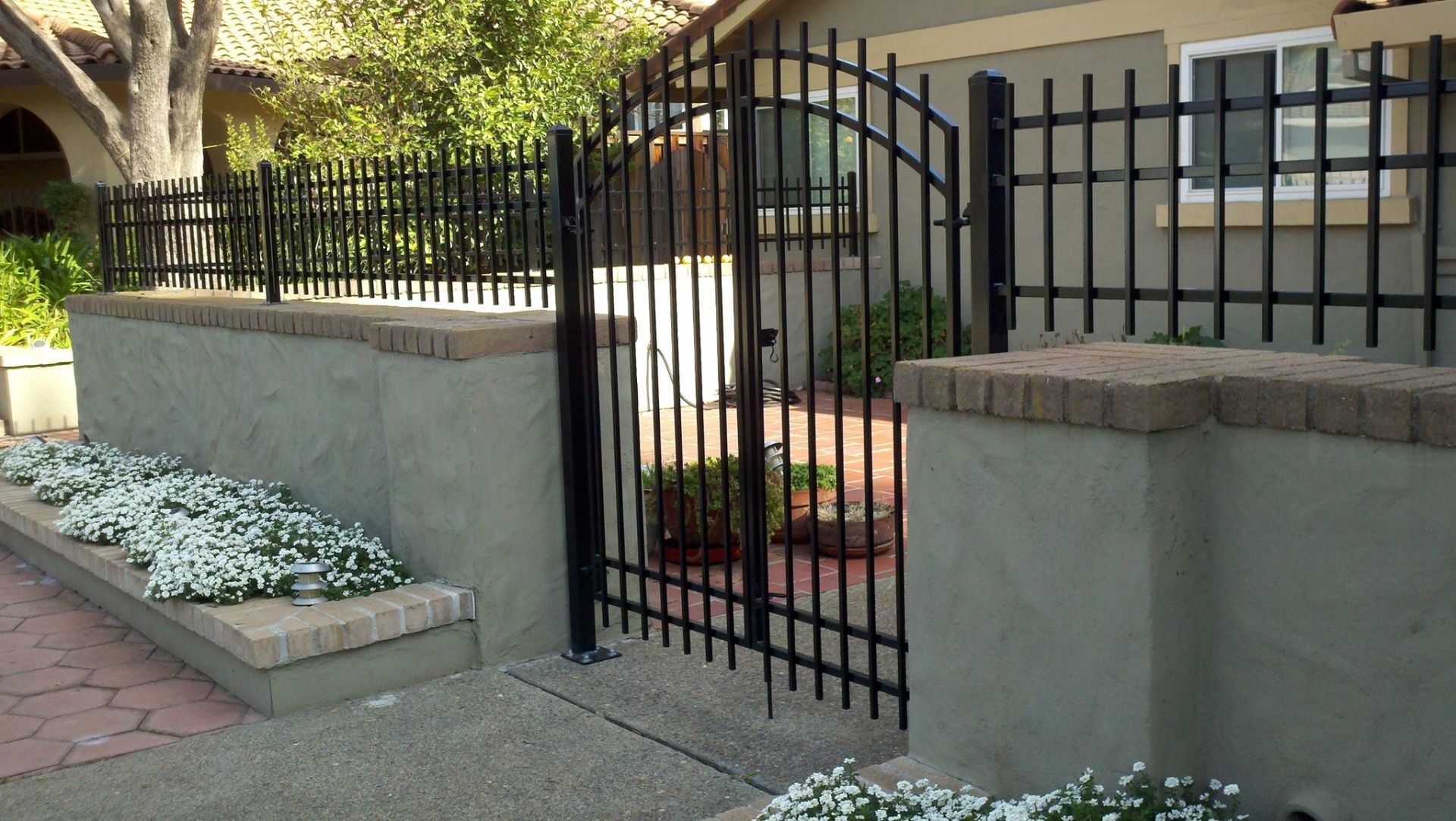 Black metal gate and fence atop a low stucco wall with brick coping. White flowers in front.