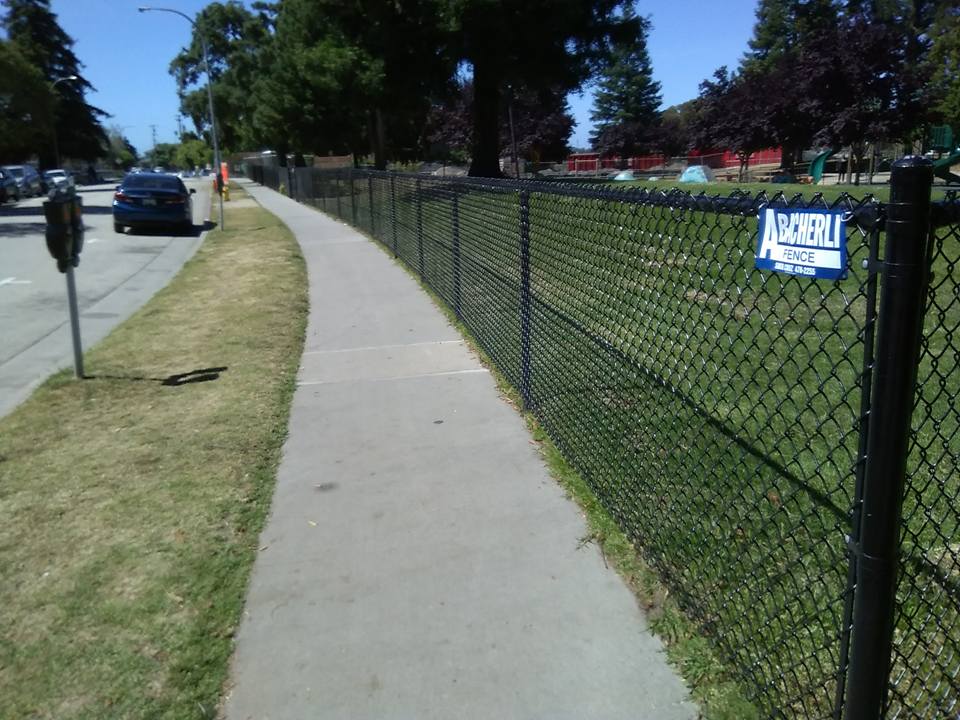 A sidewalk alongside a chain-link fence, with grass and road on either side under a blue sky.