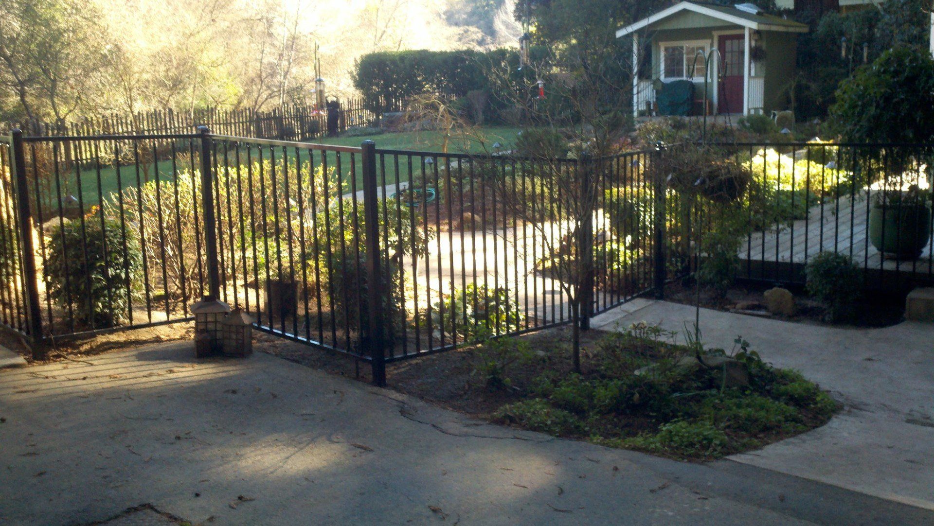 Black metal fence encloses a garden with plants and a house in the background.