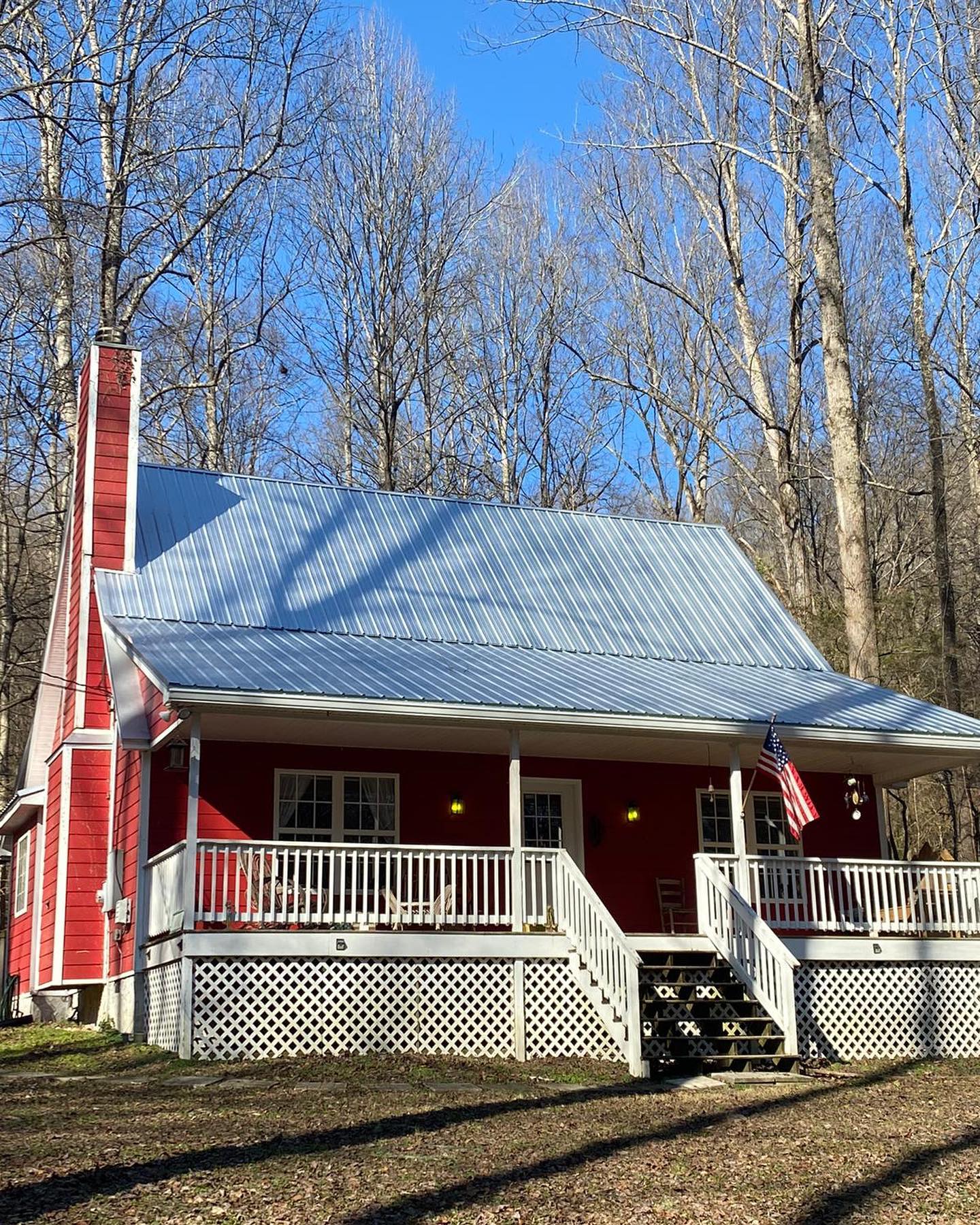 Red cabin with a gray metal roof and white porch railings, set among bare trees in daylight.
