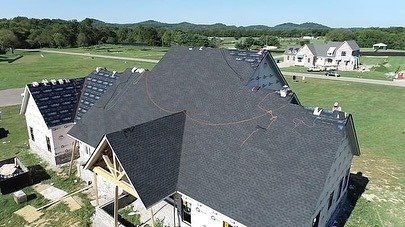 Aerial view of a large house with a dark gray roof in a green suburban neighborhood.
