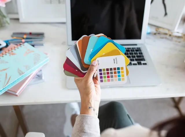 A woman is sitting at a desk holding a palette of colors in front of a laptop computer.