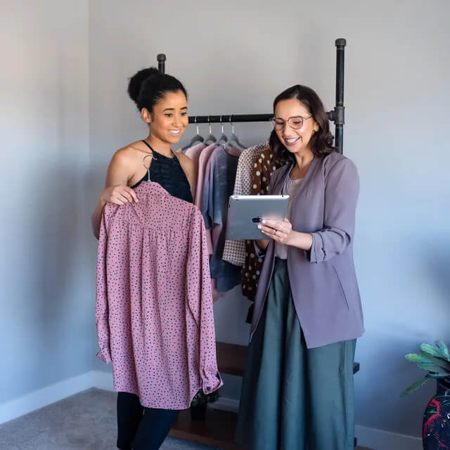 Two women are standing next to each other holding clothes and looking at a tablet.